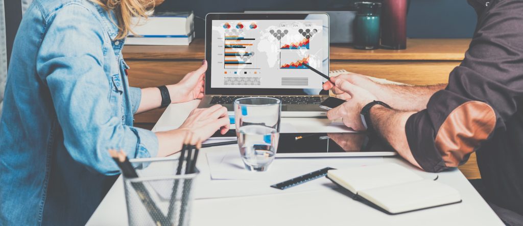 A businessman and a businesswoman are sitting at a table in front of a laptop and working on graphs, charts, and diagrams displayed on the computer screen. They appear to be discussing third-party endorsements and digital marketing.